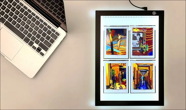 A laptop and a lightbox with four paintings on it.